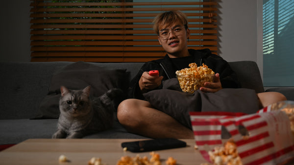 Glücklicher junger asiatischer Mann, der fernsieht und Popcorn isst, während er mit einer schönen Katze auf einem gemütlichen Sofa sitzt.