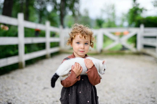 Kleines Mädchen mit Katze steht auf dem Bauernhof und blickt in die Kamera.
